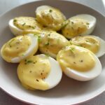 Close-up of halved boiled eggs generously topped with creamy, yellow mustard sauce and sprinkled with chives for Boiled Eggs with Mustard Sauce.