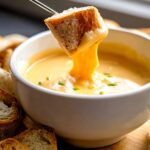A piece of crusty bread being dipped into a white bowl of smooth, melted Cheddar Cheese Fondue.