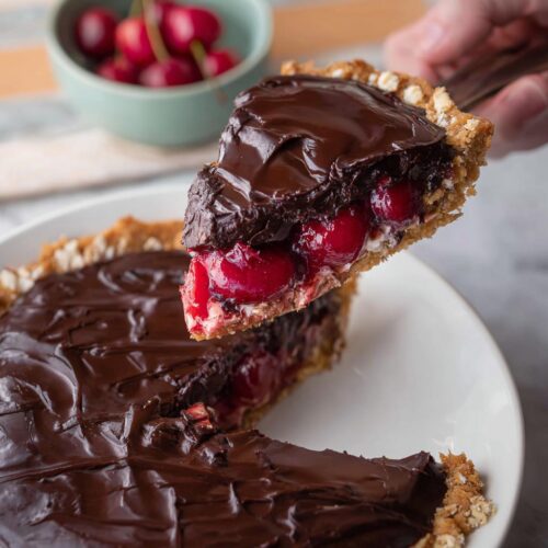 A slice of Cherry Chocolate Ganache Pie with a thick chocolate topping and bright red cherries being lifted from the rest of the decadent pie.