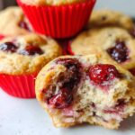 Close-up of a soft Cherry Yogurt Muffin with a bite taken out, revealing juicy cherries inside red liners.