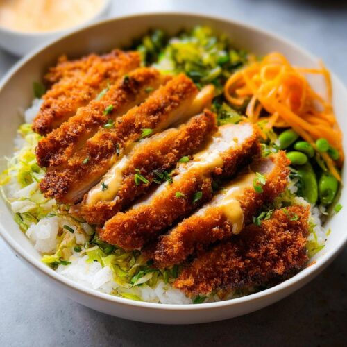 Close-up of a Chicken Katsu Bowl featuring sliced, crispy fried chicken over rice and shredded lettuce, topped with sauce and pickled carrots.