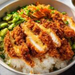 A close-up of a Chicken Katsu Bowl featuring sliced, crispy fried chicken over rice and topped with pickled vegetables.