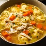 Close-up of a hearty bowl of Chicken Tortellini Dinner Soup with shredded chicken, carrots, and herbs.