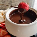 A strawberry being dipped into rich Chocolate Fondue with Heavy Cream using a fondue fork.