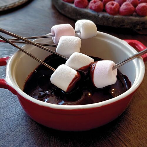Marshmallows on skewers being dipped into rich, melted chocolate fondue in a small red pot.