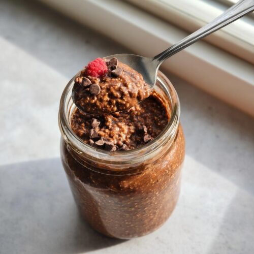 A spoonful of rich Chocolate High-Protein Overnight Oats topped with a raspberry and chocolate chips, lifted from a glass jar.