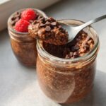 A spoonful of thick Chocolate High-Protein Overnight Oats topped with chocolate chips, with a second jar garnished with raspberries in the background.