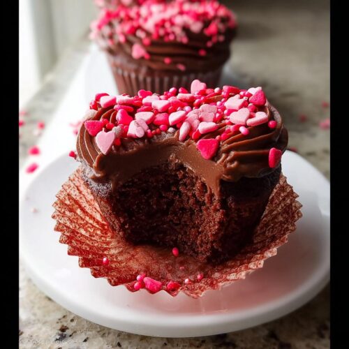 Close-up of a Chocolate Valentine Cupcake with heart sprinkles, showing a bite taken out of the rich chocolate cake.