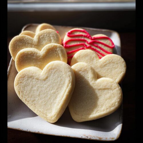 A plate of freshly baked Classic Heart-Shaped Sugar Cookies, some plain and one decorated with red icing.