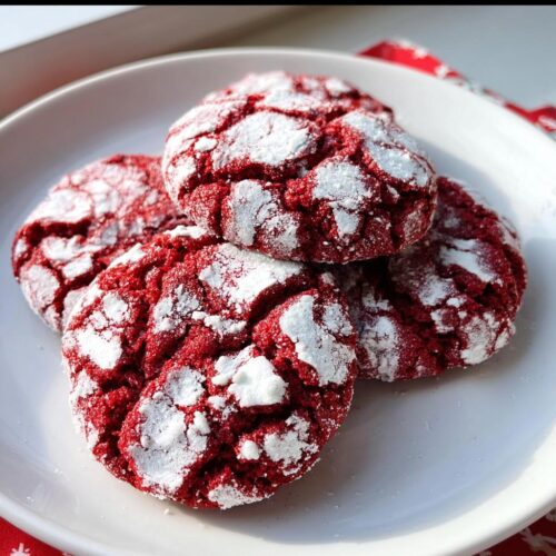 Four vibrant red Classic Red Velvet Crinkle Cookies heavily dusted with white powdered sugar on a white plate.