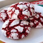 Four vibrant red velvet crinkle cookies heavily dusted with powdered sugar, resting on a white plate.