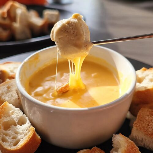 A piece of bread is dipped into a bowl of rich, melted Creamy Cheddar Cheese Fondue, showing long cheese pulls.