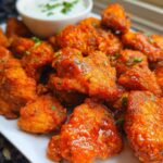 Close-up of bright orange, glazed Crispy Dragon Chicken in Air Fryer pieces piled on a white plate with a dipping sauce in the background.