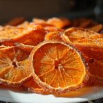 Close-up of bright orange, sugar-coated Dehydrated Candied Orange Slices piled on a white plate.
