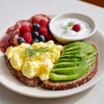 A plate featuring scrambled eggs on toast, sliced avocado, cured meat, berries, and yogurt, perfect for an Easy Breakfast for Rushed Days.