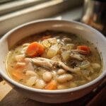 Close-up of a bowl of Family Protein Soup Dinner featuring shredded chicken, white beans, carrots, and celery in broth.