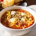 A close-up of a white bowl filled with Healthy Comfort Lasagna Soup, featuring noodles, meat sauce, and dollops of ricotta cheese.