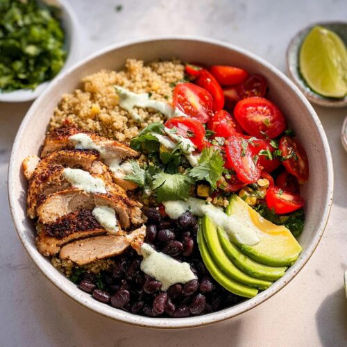 A vibrant Healthy Dinner Taco Bowl featuring sliced seasoned chicken, quinoa, black beans, cherry tomatoes, avocado, and a creamy green drizzle.