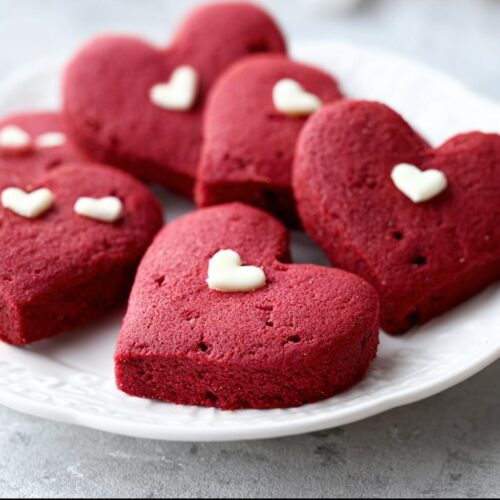 Several vibrant red, heart-shaped red velvet cookies topped with small white candy hearts on a white plate.