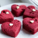 Five vibrant red, heart-shaped red velvet cookies topped with small white chocolate hearts on a white plate.