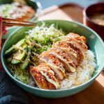 A vibrant Katsu Bowl with sliced crispy chicken, avocado, shredded cabbage, and a drizzle of sesame dressing over rice.