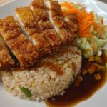 A plated Katsu Bowl with brown rice, topped with sliced, crispy fried chicken and drizzled with sauce, served with shredded vegetables.