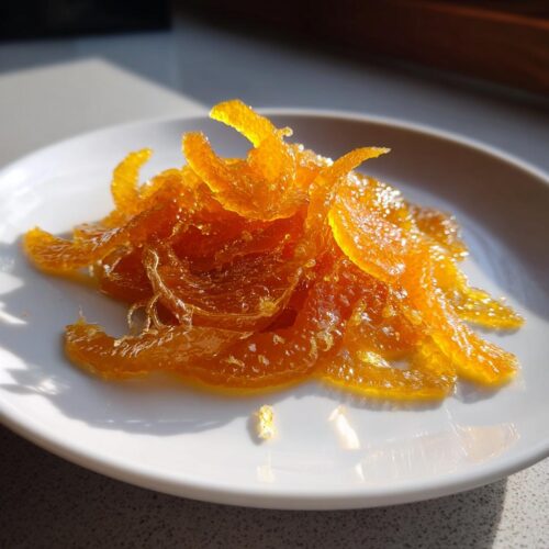 Close-up of glistening, candied orange strips, representing Mixed Peel with Vanilla Syrup, piled on a small white plate.