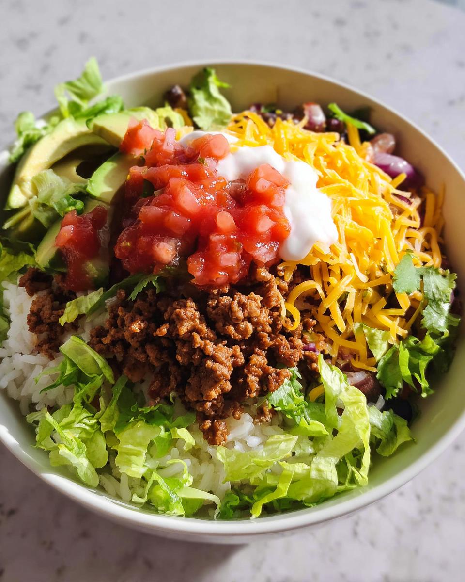 A close-up of a hearty One-Bowl Taco Dinner featuring rice, seasoned ground beef, lettuce, cheese, salsa, and sour cream.