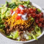 A close-up of a fully loaded One-Bowl Taco Dinner featuring rice, seasoned ground beef, lettuce, cheese, tomatoes, and sour cream.