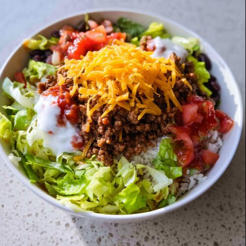 Close-up of a One-Bowl Taco Dinner featuring rice, seasoned ground beef, cheese, lettuce, salsa, and sour cream.