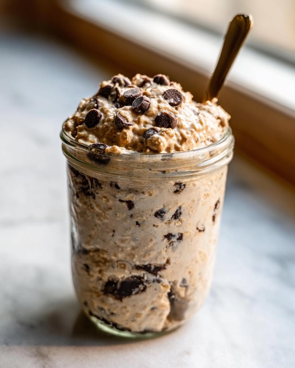 A jar filled with thick Protein Cookie Dough Overnight Oats topped with chocolate chips and a spoon.