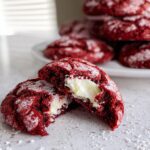 A Red Velvet Crinkle Cookie split in half showing a gooey cream cheese center, dusted with powdered sugar.