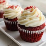 Close-up of two Red Velvet Valentine Cupcakes topped with swirled cream cheese frosting and red crumbs.