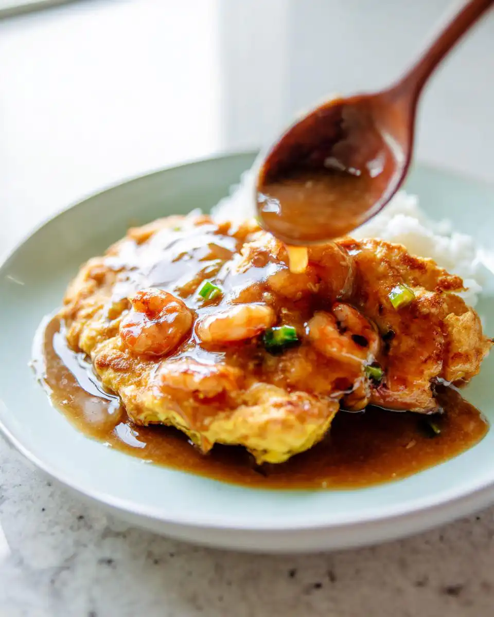 A wooden spoon pours light soy gravy over a serving of Shrimp Egg Foo Young resting on white rice.
