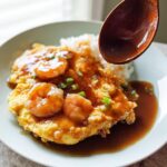 A wooden spoon pours light soy gravy over Shrimp Egg Foo Young served with white rice on a light blue plate.