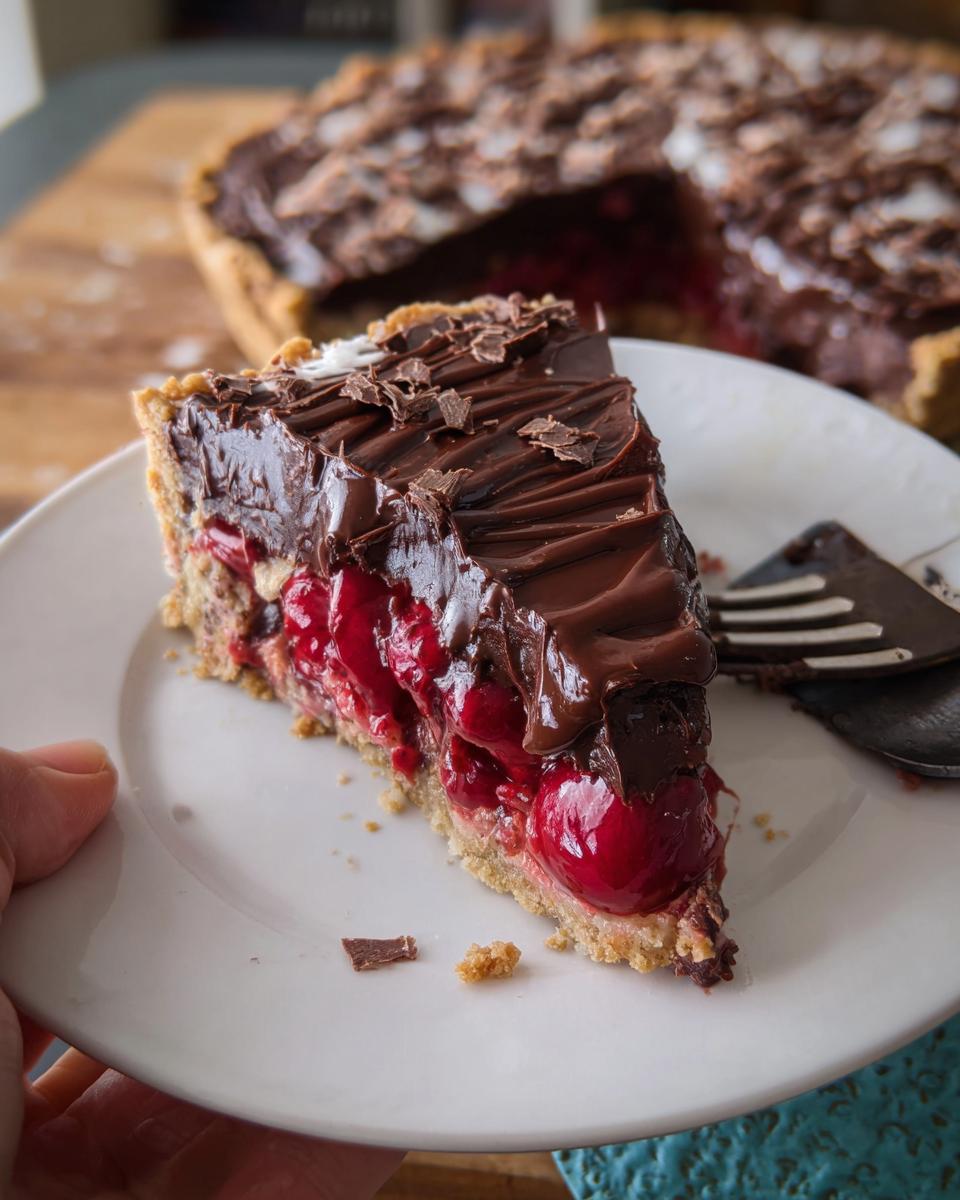 A decadent slice of Cherry Chocolate Ganache Pie showing a thick layer of whole cherries beneath rich chocolate ganache and shavings.