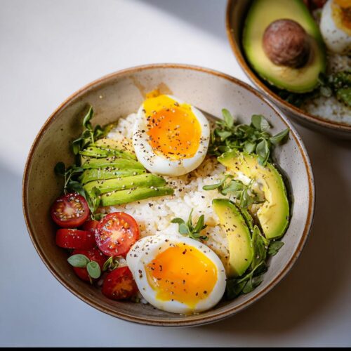 Two halves of soft Boiled Eggs for Breakfast Bowls served over rice with avocado slices and cherry tomatoes.