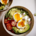 Close-up of a breakfast bowl featuring soft boiled eggs for breakfast bowls, sliced avocado, and tomatoes on rice.