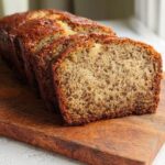 A loaf of freshly baked Soft & Moist Banana Bread, partially sliced, resting on a rustic wooden cutting board.