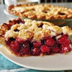 A close-up slice of Sour Cherry Almond Pie showing juicy filling and flaky crust, with the whole pie blurred in the background.