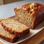 A golden-brown Sour Cream Banana Cake Loaf, partially sliced, resting on a white platter.