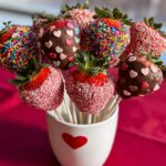 A bouquet of chocolate-dipped strawberries decorated with colorful sprinkles and pink heart candies, ready for presentation.