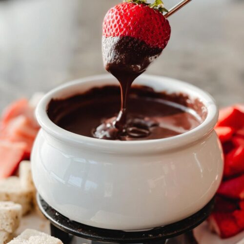 A fresh strawberry being dipped into rich Chocolate Fondue with Heavy Cream using a fondue fork.