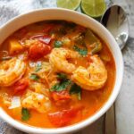A close-up, overhead view of a white bowl filled with vibrant Thai Red Curry Shrimp Soup, featuring large shrimp, tomatoes, and cilantro.