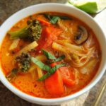 Close-up of a bowl of vibrant Thai Red Curry Vegetable Soup, featuring broccoli, carrots, and basil.