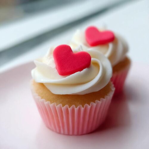 Two vanilla Valentine Cupcakes with white frosting and bright red sugar heart toppers, sitting on a pale pink plate.