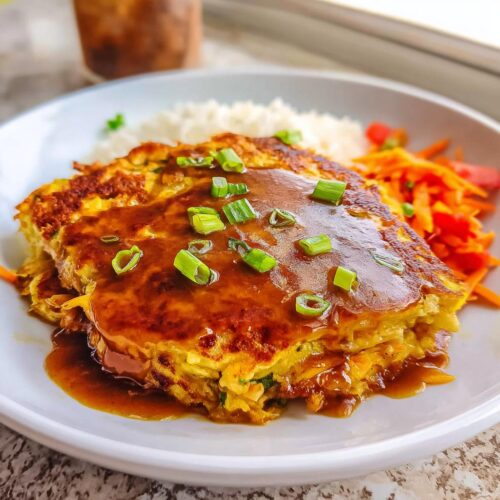 A serving of Vegetable Egg Foo Young smothered in brown gravy and topped with chopped green onions, served with rice.