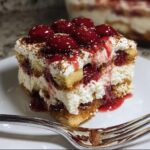 A decadent slice of White Chocolate Cherry Tiramisu topped with whole cherries and chocolate dust, resting on a white plate.