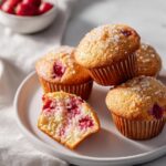A stack of golden Bakery Style Strawberry Muffins topped with coarse sugar, one cut open showing berries.