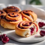 A close-up view of freshly baked Cherry Brioche Rolls, one cut open to show the filling, garnished with fresh cherries.